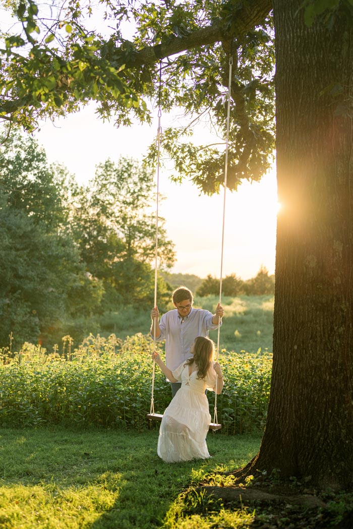 Engaged couple posing on a tree swing in front of a flower field at Firefly Haven in Hamptonville, NC. Photo by Ashley Logan Photography.